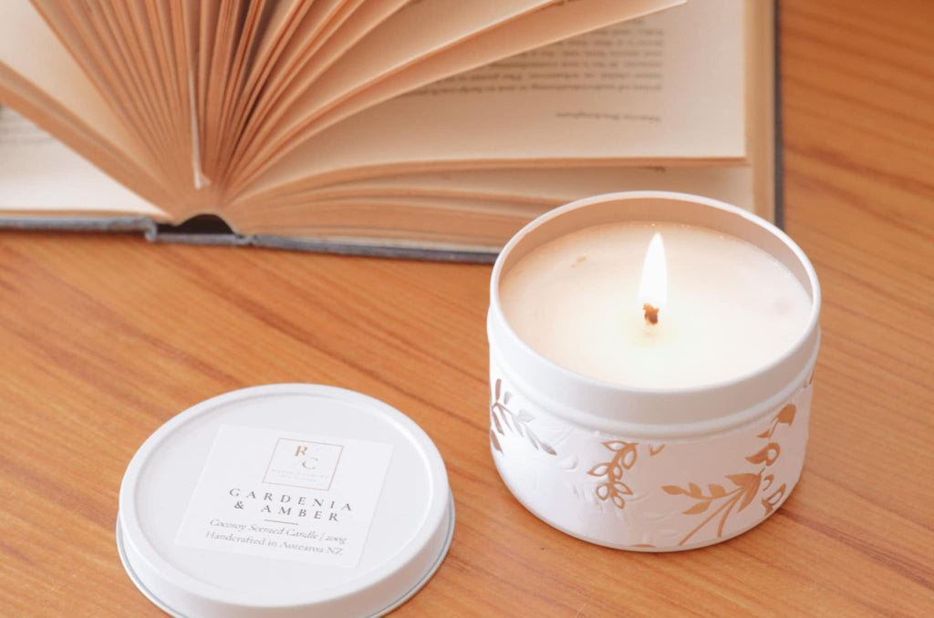 A lit white candle in a white tin with gold botanical illustrations sits on a wooden table next to its lid, with an open book blurred in the background. The label reads 'GARDENIA & AMBER'.